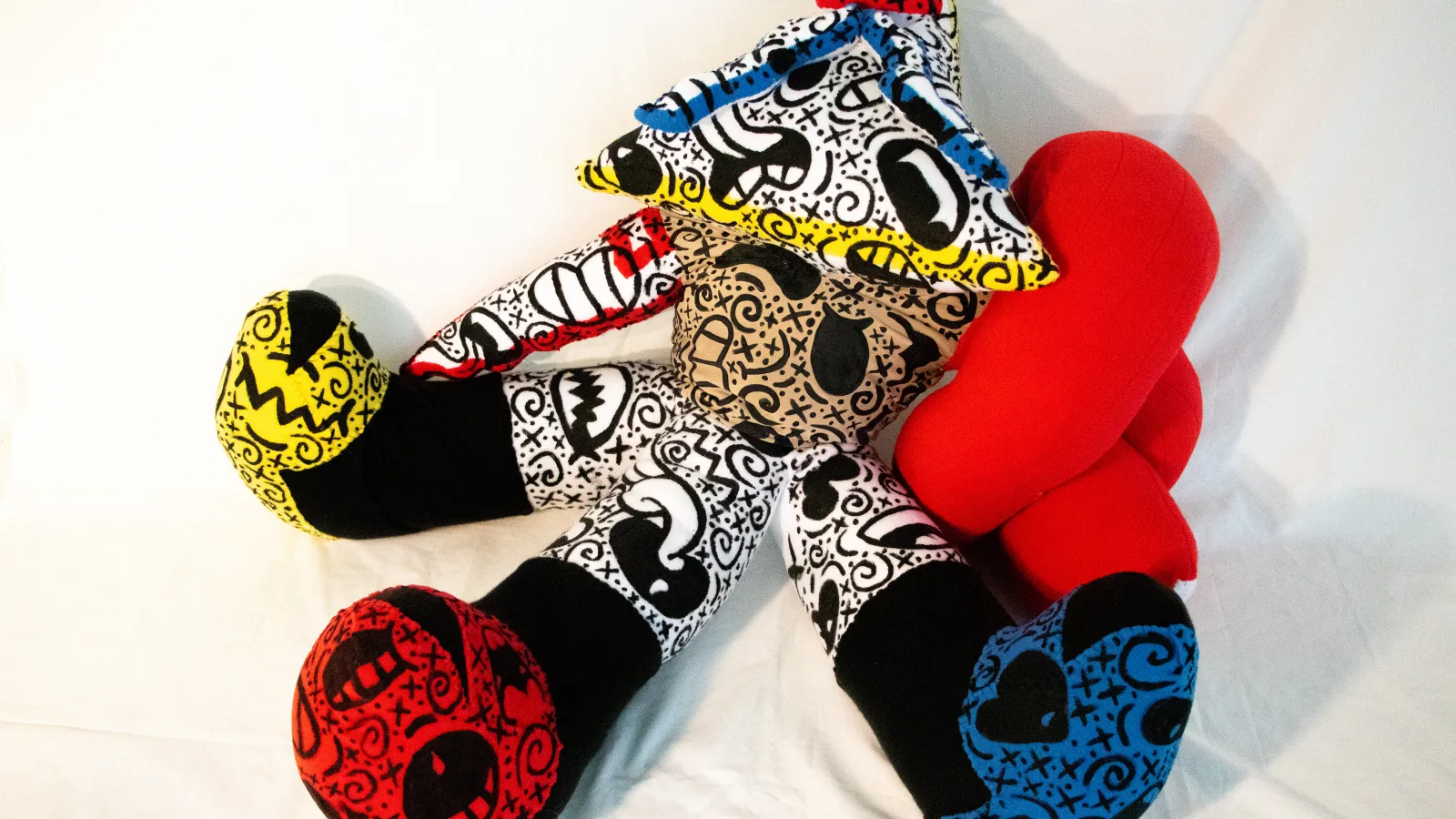 A photograph of a fabric sculpture in bright red, yellow and blue with multiple(3) appendages and covered with swirling black markings, save for one purely red portion and three entirely black pieces near the end of the appendages.. 