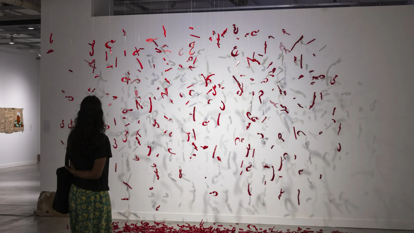 visitor looking at sculptural display of Illya's work--red letters in Persian/Farsi hanging from the ceiling in a deconstructed poem