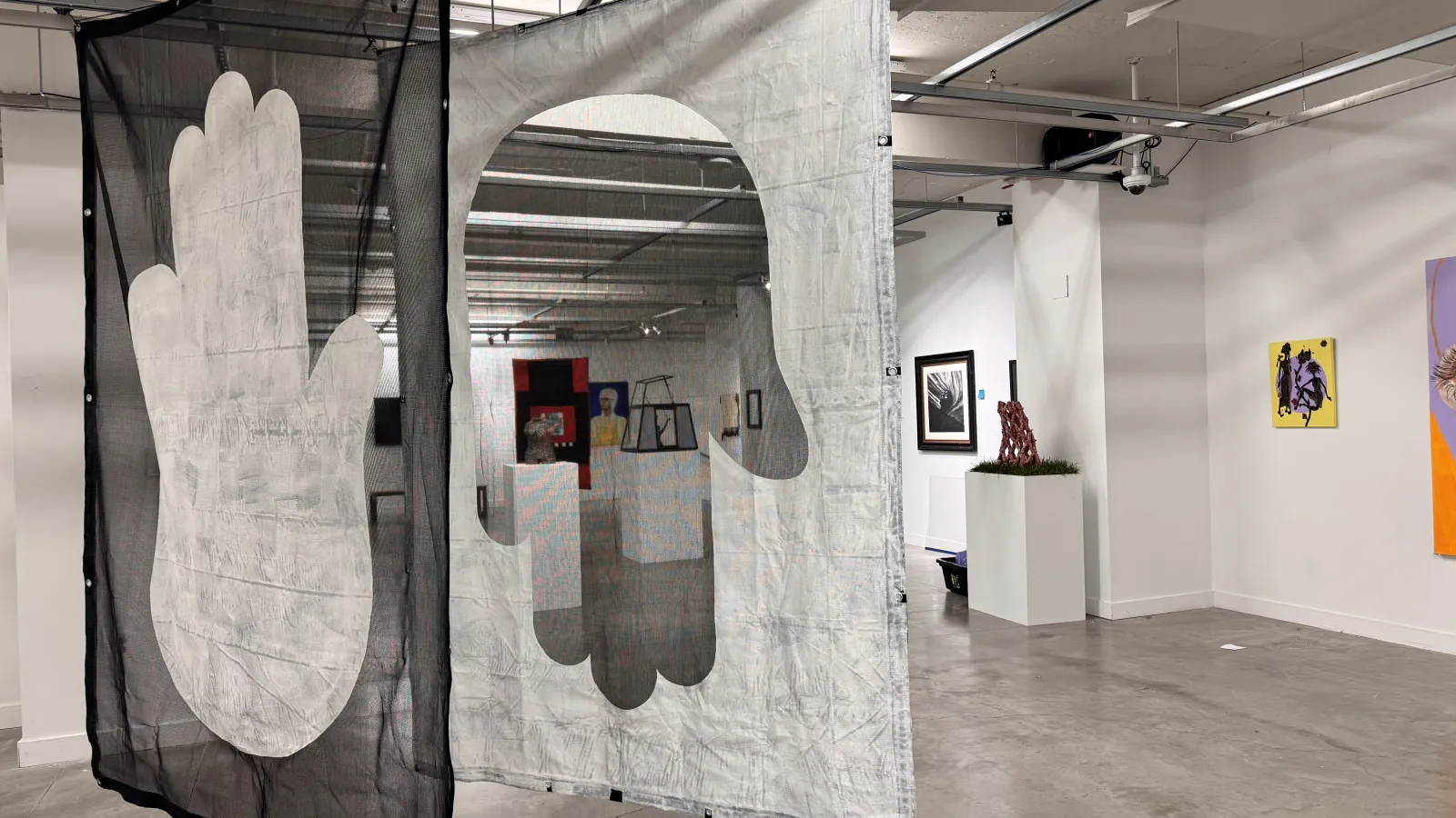 Installation view of an art gallery featuring two large hanging fabric panels with the outline of a hand printed on them, dividing the space. The panel on the left is black with a white hand, and the one on the right is white with a cutout of a dark hand, creating a layered effect. In the background, other paintings and sculptures are visible on white walls.