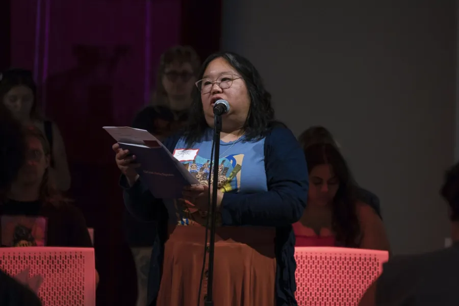 A person in a long skirt standing at a microphone reading from a paper