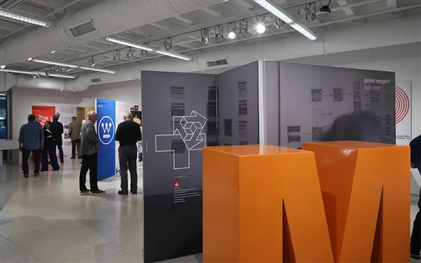 Gallery space with a large orange M and people looking at artwork. 