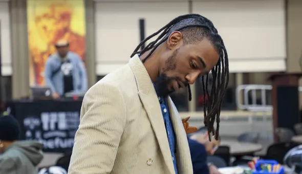 a man of color with long hair and in a tan suit looking down to the ground