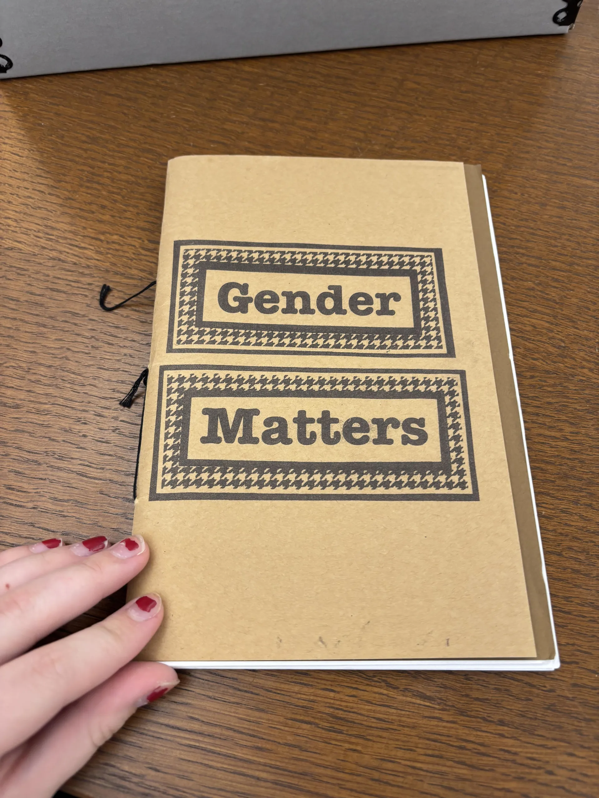 Zine titled Gender Matters on a table with a brown cardstock cover and the words in houndstooth boxes. The zine is bound with string.